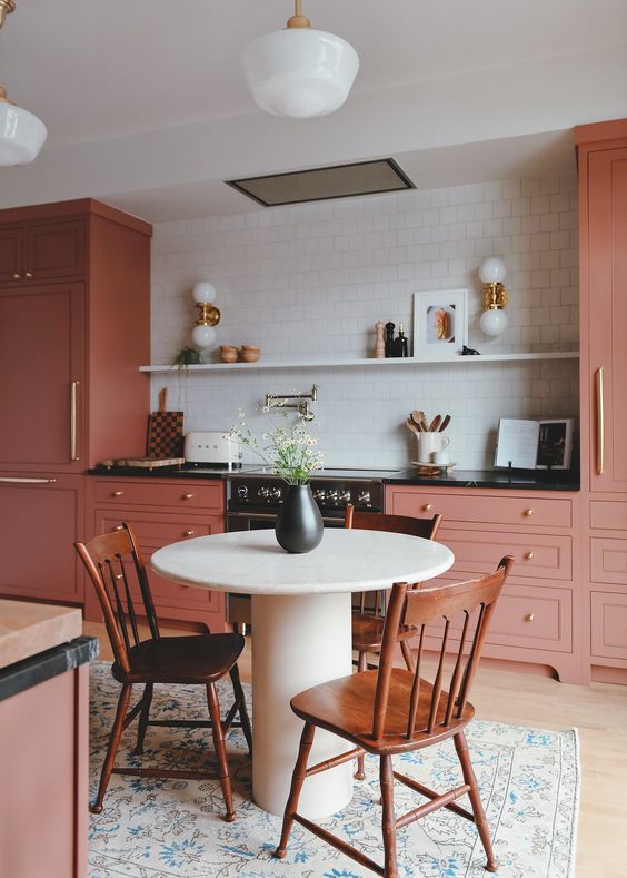 a pink kitchen with a black countertop, a backsplash made of white square tiles, an open shelf with decor and pretty sconces