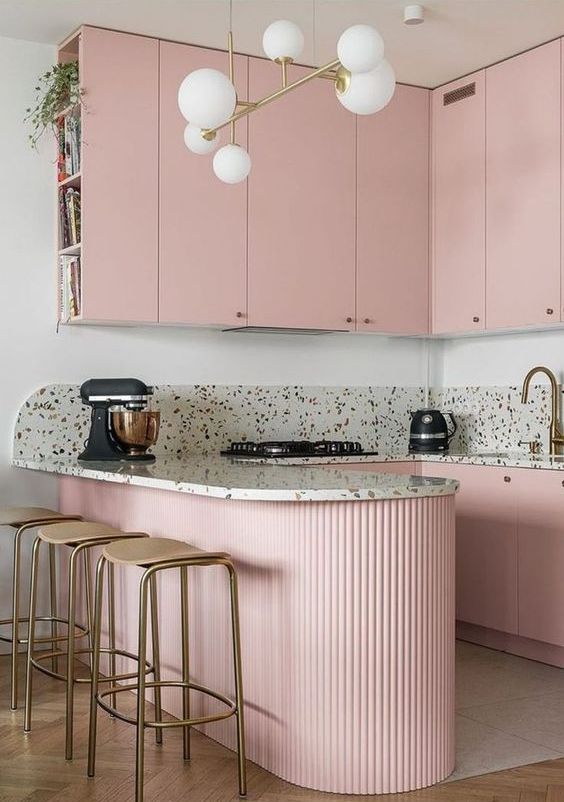A pretty pink kitchen with sleek upper cabinets and a fluted kitchen island, terrazzo countertops and a gold accent backsplash