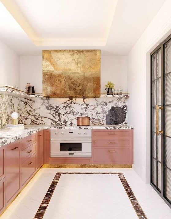A sophisticated and glamorous kitchen with pink cabinets, a marble backsplash and countertops and a gold extractor hood looks stunning