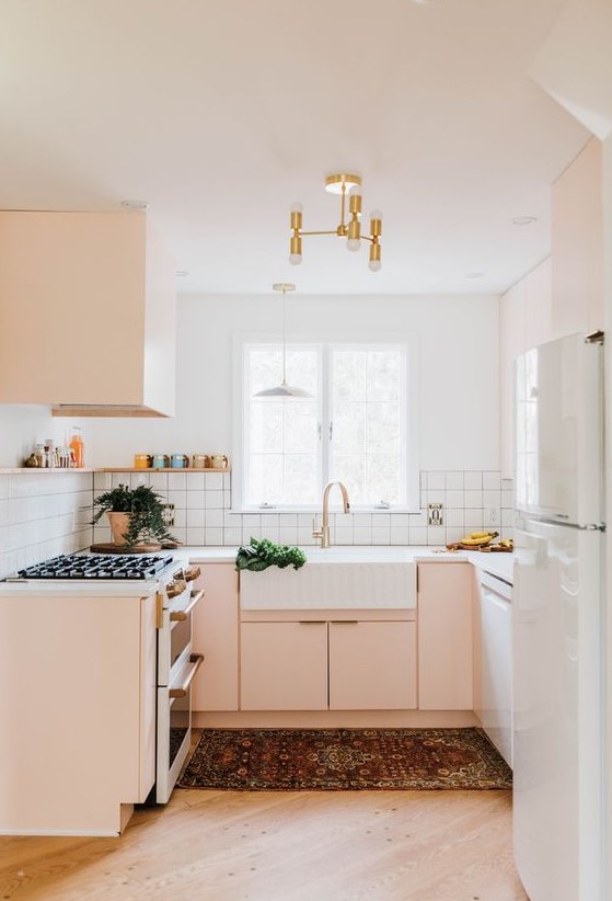 A small but chic blush and white kitchen with a gold chandelier, white tile backsplash and white countertops