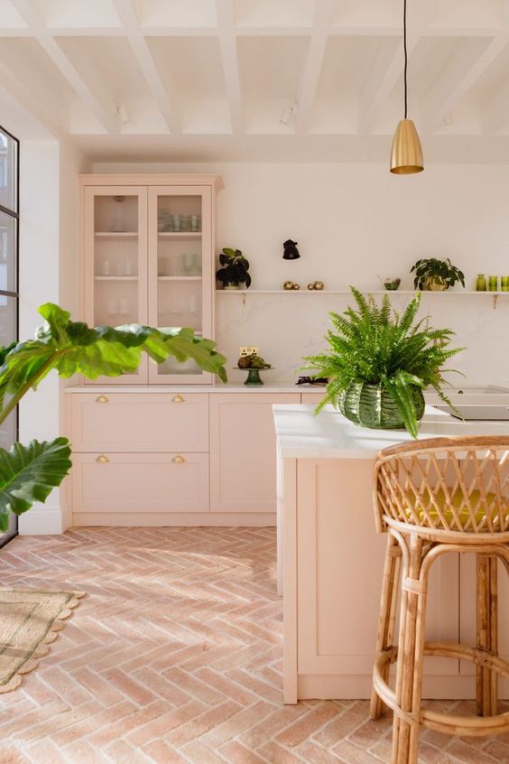 an inviting pink kitchen with shaker cabinets, a large kitchen island, white stone countertops, a mantel with potted plants and rattan stools