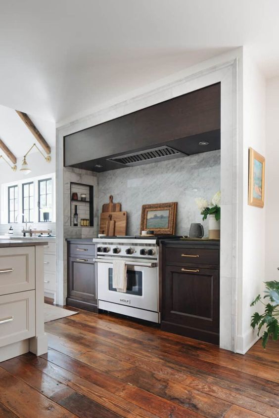 A modern farmhouse kitchen in white with a large alcove with dark stained cabinets and an extractor hood as well as some small shelves for spices and oils