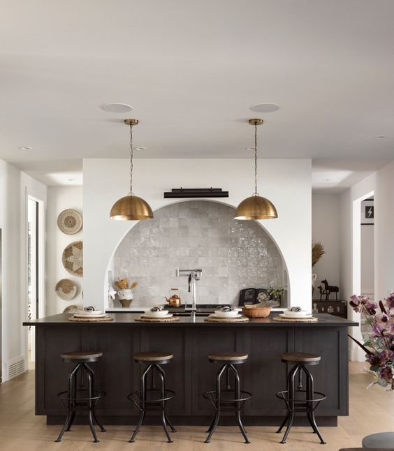 a modern kitchen with an arched zellige-tiled alcove and built-in cabinets, a black island, brass pendant lamps and industrial stools