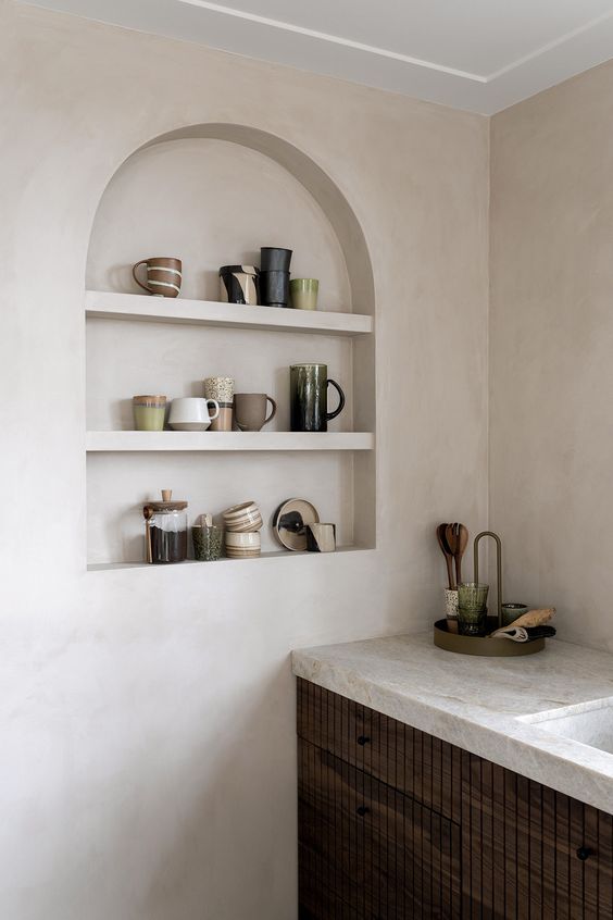 a modern kitchen with plaster walls, dark stained cabinets and neutral stone countertops, an arched niche for displaying cups