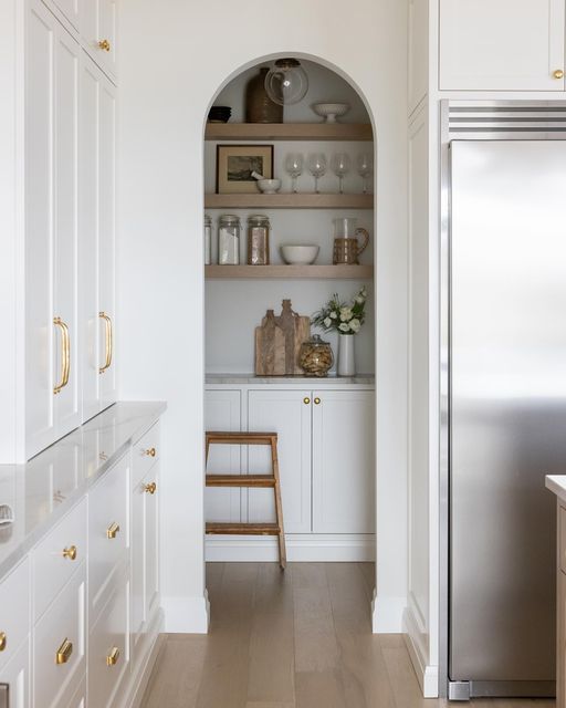 A white, modern farmhouse kitchen with an arched nook that hides a pantry with cabinets and open shelves - a clever solution to save some time