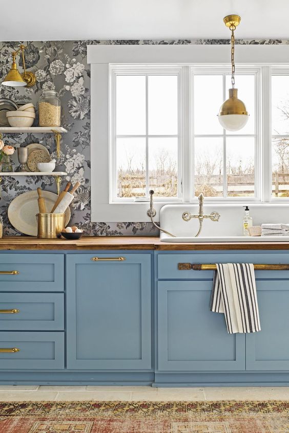 a sophisticated kitchen with gray floral print wallpaper, blue shaker cabinets, butcher block countertops, gold handles and a gold pendant lamp