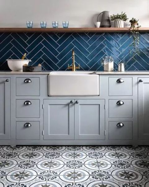 a stylish kitchen with dove gray cabinets, a navy herringbone tile backsplash, open shelving and printed tiles on the floor