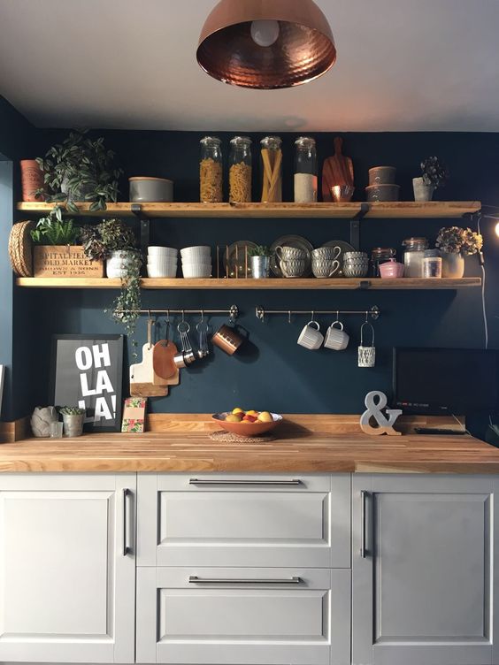 A dove gray kitchen with a navy blue wall, wooden shelves and butcher block countertops, and touches of copper and green