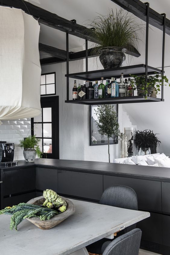 a black industrial kitchen with elegant cabinets, a hanging shelf above the kitchen island, black beams and lots of greenery around it