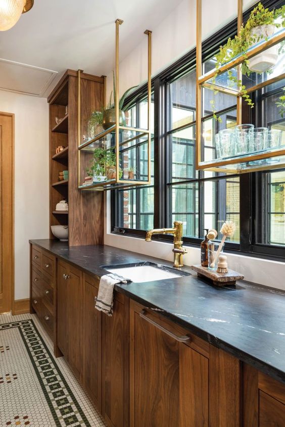A stained kitchen with black stone countertops and hanging brass shelves above the windows is a cool and elegant space
