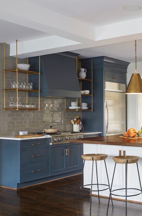 A sleek blue kitchen with a glossy gray tile backsplash, hanging brass shelves, brass pendant lamps and stools