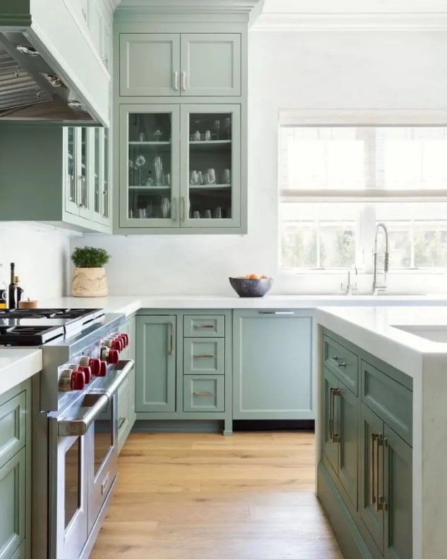 A beautiful sage green kitchen with shaker and glass front cabinets, white stone countertops and a backsplash feels airy