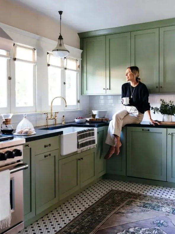 A sage green farmhouse kitchen with shaker cabinets, black countertops, a white subway tile backsplash, and brass handles