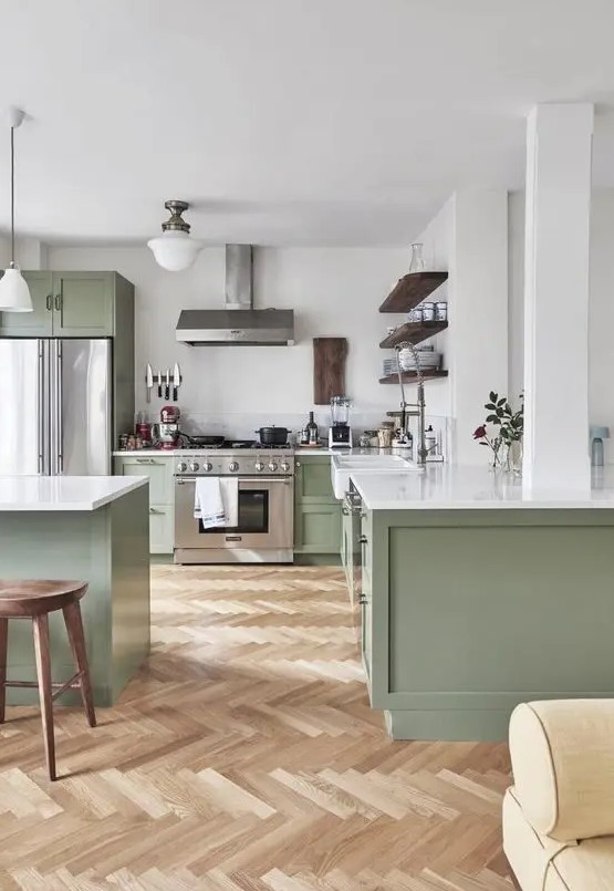 A large sage green kitchen with shaker cabinets, white stone countertops, open shelving, and wooden stools is sleek and cool