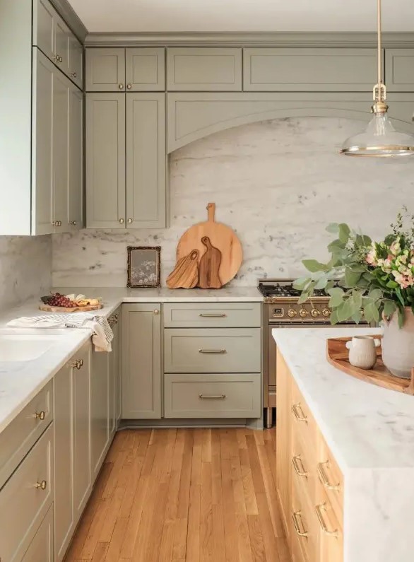 A beautiful sage green kitchen with shaker cabinets, a white stone backsplash and countertops, a stained kitchen island, and a chic pendant lamp
