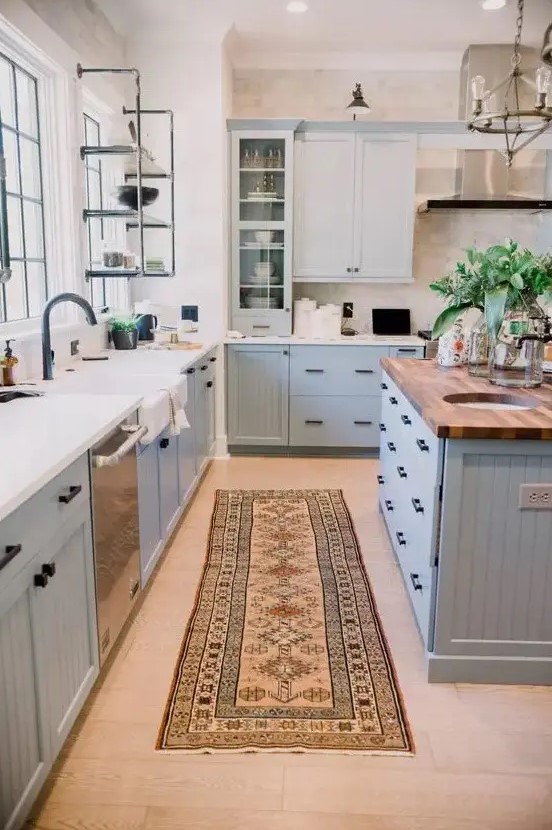 A modern country kitchen in light blue with shaker style cabinets, white and butcher block countertops, open metal shelving and a metal hood