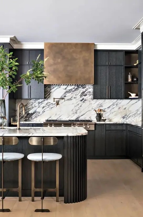 a sleek black kitchen with a white marble backsplash, an aged brass range hood, a fluted curved kitchen island and tall brass-backed stools
