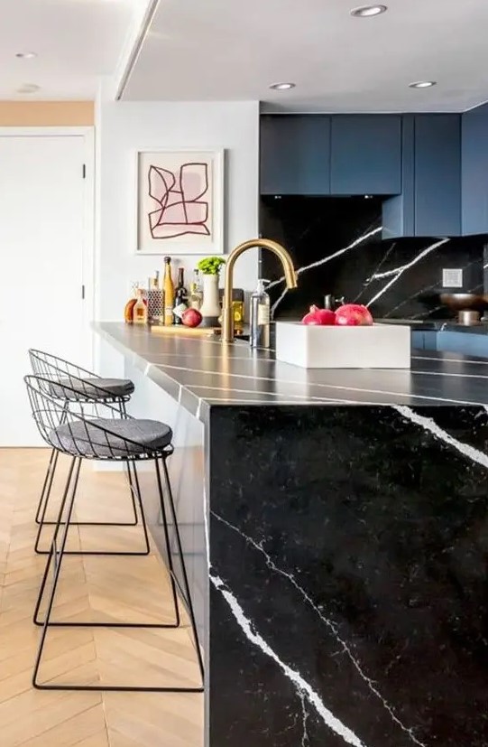 A blue kitchen with a black marble backsplash and a waterfall black countertop and touches of gold for a chic look