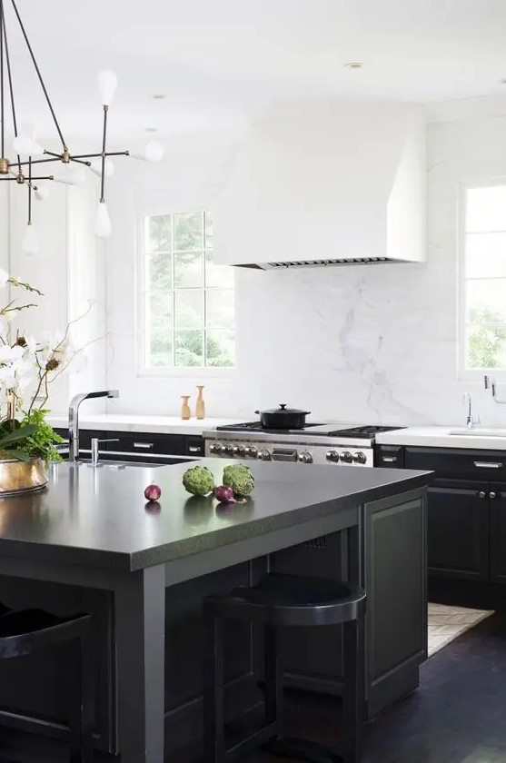 a cool black kitchen with a large white extractor hood, a large black kitchen island and matching stools, a chandelier and some brass elements