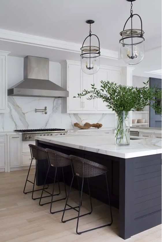 A modern farmhouse kitchen with white shaker-style cabinets, a large metal hood, a black island, a white marble backsplash and countertops