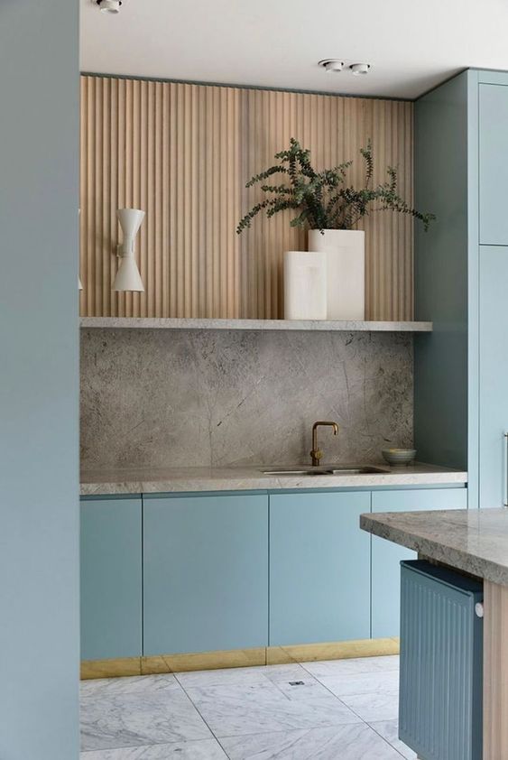 a minimalist kitchen with light blue and stained fluted cabinets, gray stone countertops and a backsplash, and potted plants