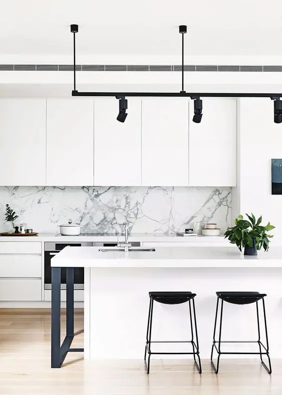a modern white kitchen with a marble backsplash, black stools, lamps and legs, and potted plants that refresh the space