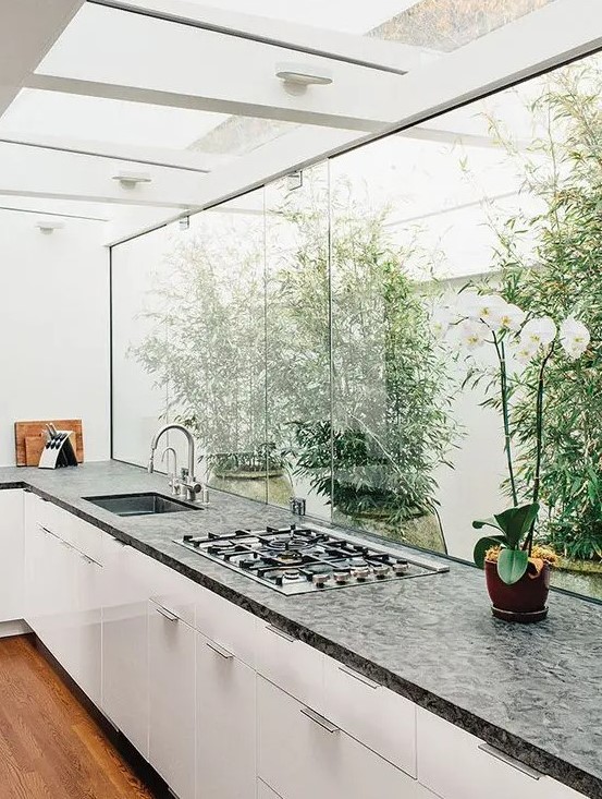 a modern white kitchen with dark worktop, a glazed wall overlooking the private garden and greenery