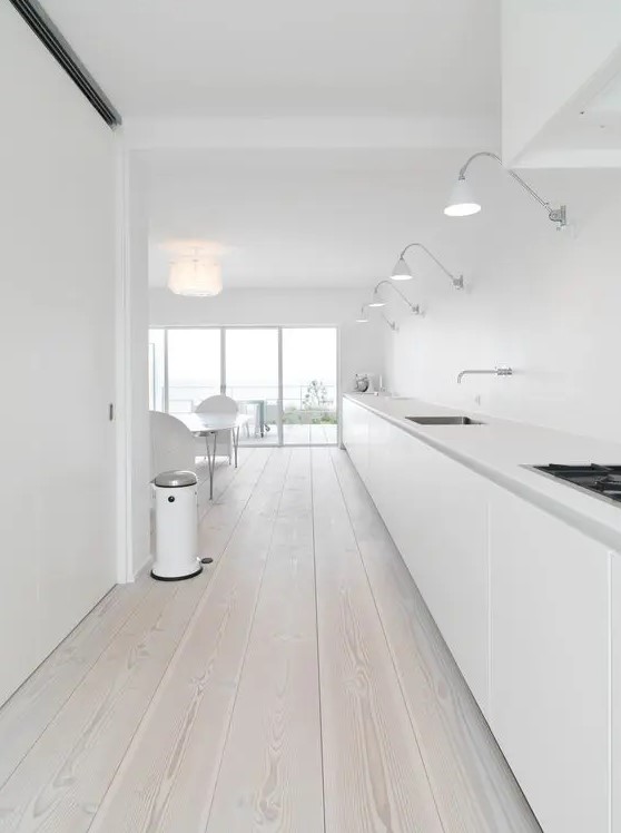 a large and super long minimalist white kitchen with just base cabinets and whitewashed floors and lights above the cabinets