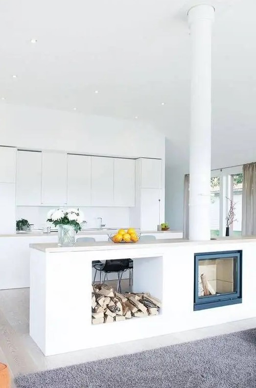 A minimalist white kitchen is made more interesting by adding a kitchen island with a built-in fireplace and firewood storage
