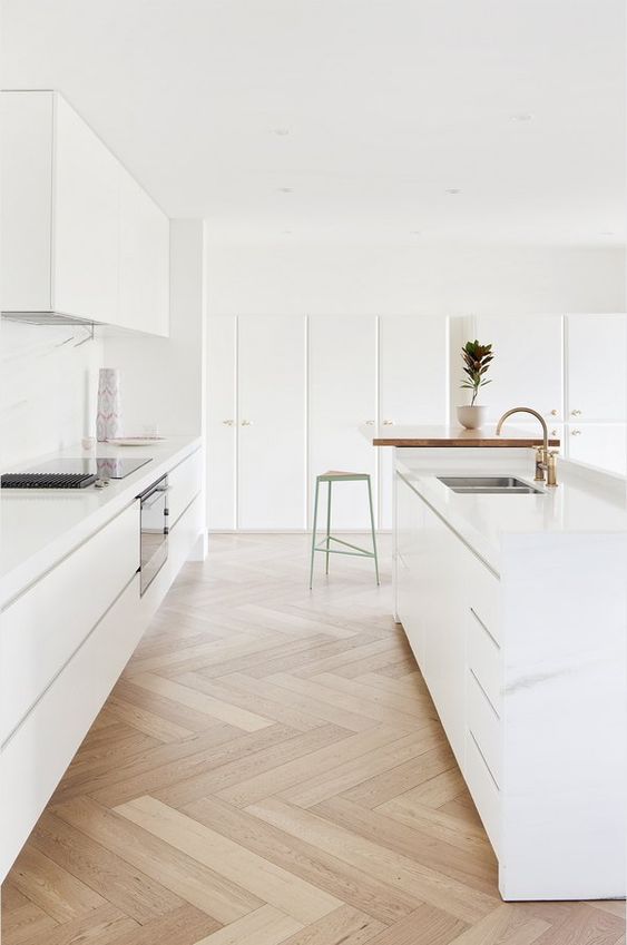 a minimalist white kitchen with sleek cabinets, a large island, white countertops and backsplash, and gold fixtures