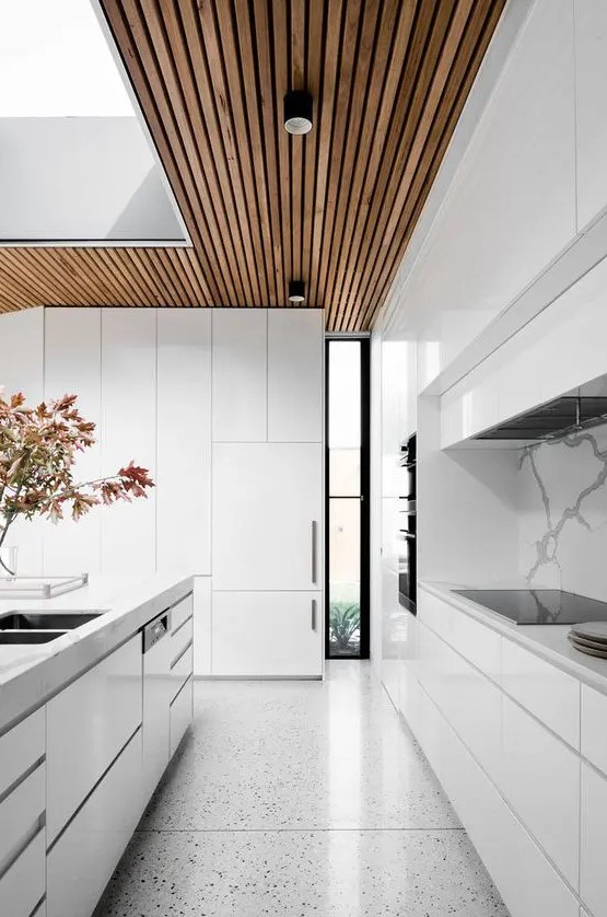 a minimalist white kitchen with sleek cabinets, a wooden ceiling and terrazzo floors, and a large skylight to maximize natural light