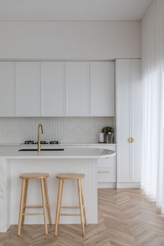 a sophisticated white kitchen with shaker-style cabinets, a slat backsplash and island, stained stools and brass handles