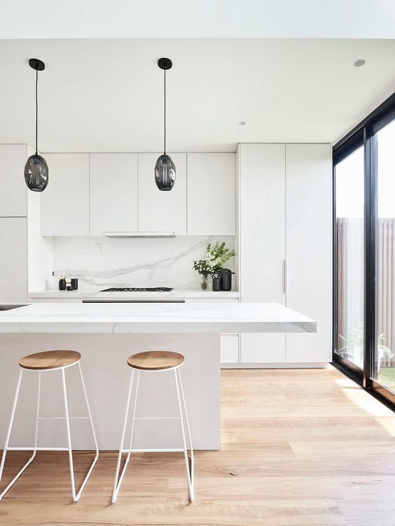 a sleek, minimalist white kitchen with a white marble backsplash, black pendant lamps above the kitchen island, and high stools
