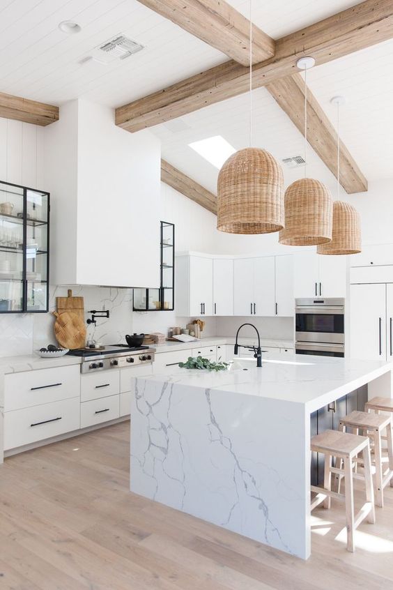 a white modern kitchen with white cabinets and black handles, a large extractor hood, a kitchen island with a seating area and woven pendant lamps