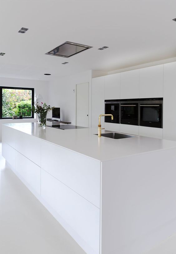 a white, ultra-minimalist kitchen with sleek cabinets, an oversized island and built-in appliances, and gold fixtures