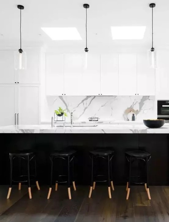 a modern kitchen with white marble countertops and backsplash, white shaker cabinets with minimalist black handles, and glass pendant lights with black cords