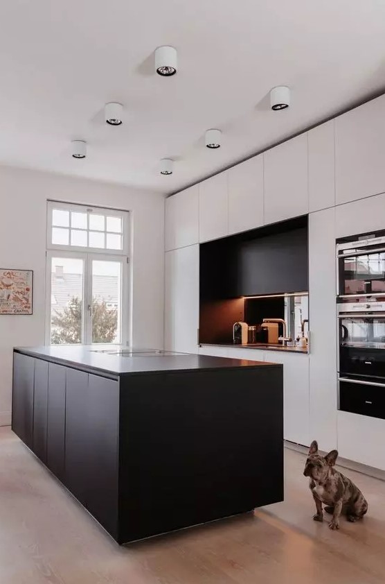 a minimalist kitchen with white cabinets and a black kitchen island, a black additional cabinet and a mirrored back wall and white lamps