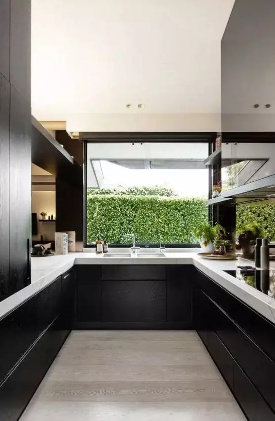Black wooden cabinets pair beautifully with white marble countertops to create a perfect modern kitchen, and a window overlooking the greenery fills the room with light