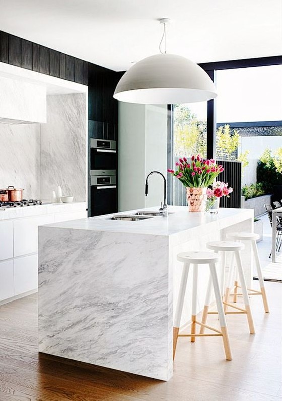 A chic white kitchen with white stone countertops and a splashback and a waterfall countertop on the kitchen island