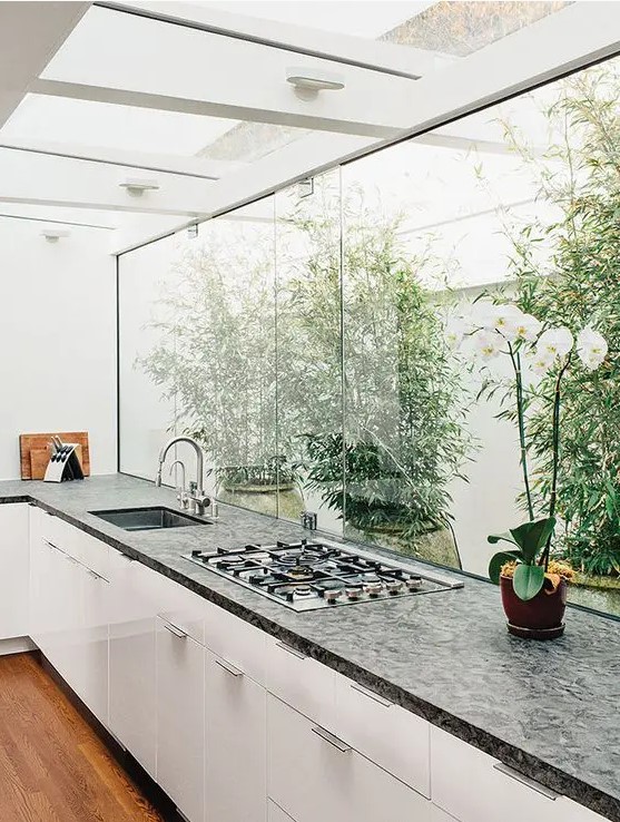 a modern white kitchen with dark worktop, a glazed wall overlooking the private garden and greenery