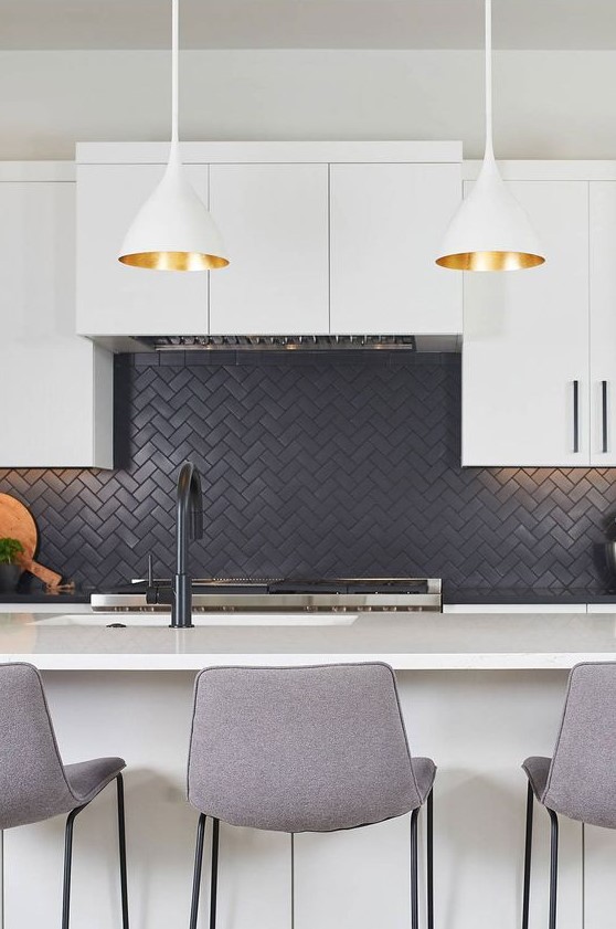 A modern white kitchen with a black herringbone backsplash, tall gray stools and pendant lamps with gold interior