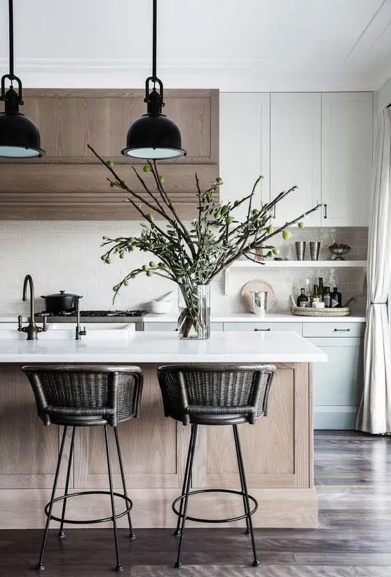 A gorgeous modern kitchen with white dove gray shaker cabinets, a stained range hood, a stained kitchen island, black pendant lamps and woven stools