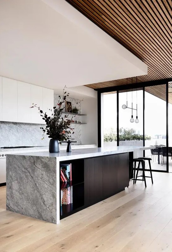 a beautiful modern kitchen with sleek white cabinets, a gray stone backsplash and countertops, a black island and a tiered ceiling