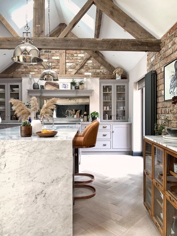 a barn kitchen with brick walls, gray Shaker-style cabinets, an elegant stone kitchen island with rusty stools and old wooden beams