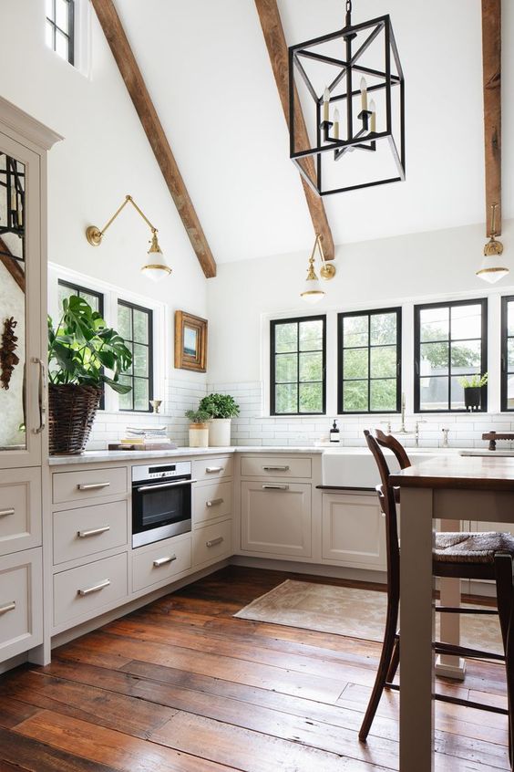 a light-filled barn kitchen with exposed beams, gilded sconces, neutral cabinets, white stone countertops, and a table serving as a kitchen island