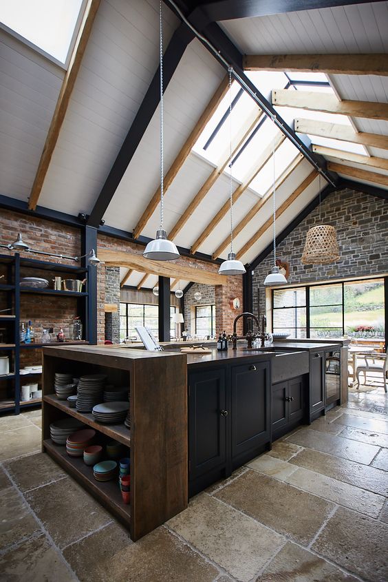 a moody kitchen with skylights, pendant lamps highlighting the double-height ceiling, dark cabinets, a wooden console table and stone walls for structure