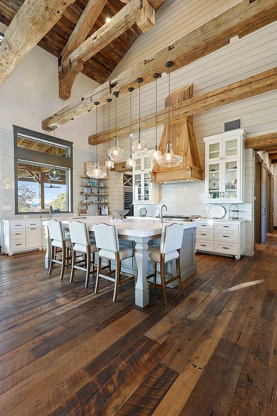 a rustic barn kitchen with light stained wood beams and a matching range hood, a white kitchen island with high stools, white cabinets and an array of hanging lamps
