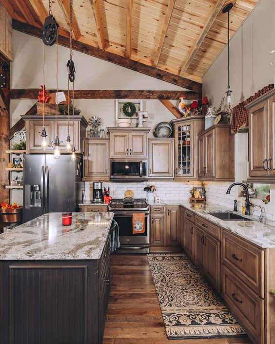 a rustic barn kitchen with exposed beams, stained cabinets, a dark-stained island, stone countertops and pendant lamps