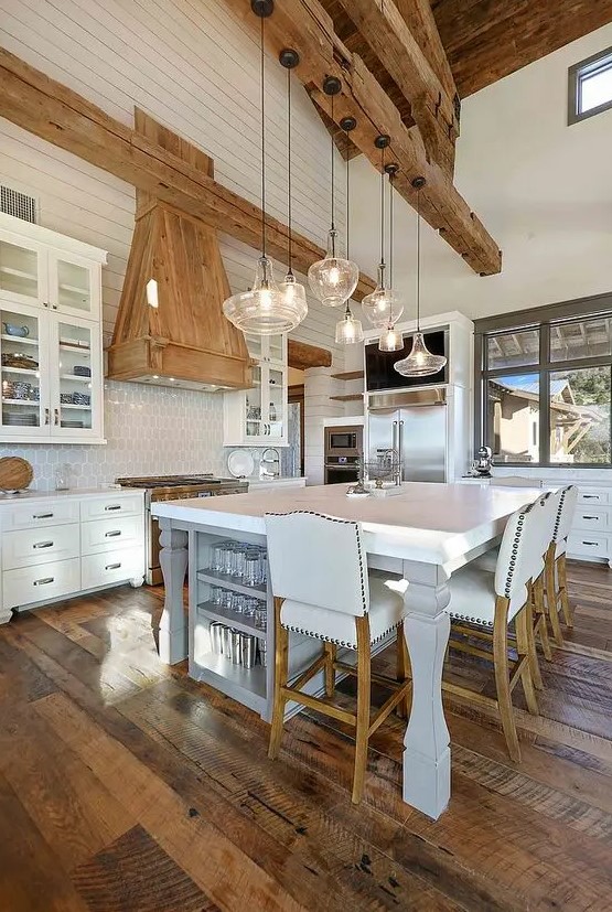 a white barn kitchen with a board ceiling, wooden beams, white cabinets and a large kitchen island that doubles as a table
