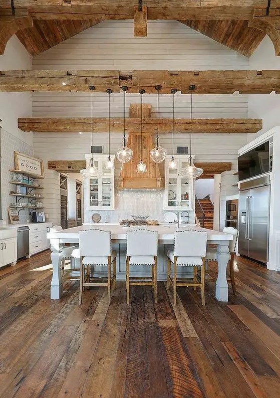an elegant white barn kitchen with board walls, stained wood beams, shaker-style cabinets, a vintage kitchen island and white stools